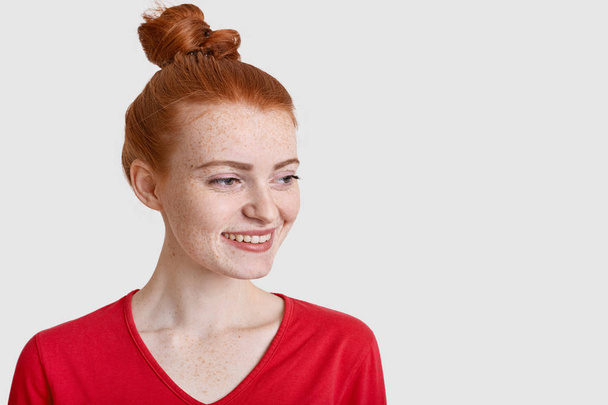 Photo of glad teenage girl with satisfied expression, focused aside, has positive outlook, thinks about something pleasant, wears red jumper isolated over white studio wall with blank space for advert - Photo, Image