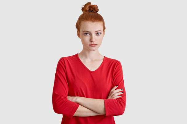 Studio shot of pleasant looking serious woman has stern expression, keeps arms folded, has red hair, listens something attentively isolated over white background. Redhaired female student poses indoor - Photo, Image
