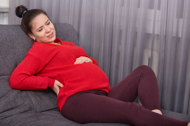 Φωτογραφία: δυσαρεστημένοι έγκυο γυναίκα αισθάνεται πόνο στην πλάτη, συνοφρυώματα πρόσωπο από δυσαρέσκεια, φοράει casual ρούχα, έχει προβλήματα με την υγεία, κάθεται στο άνετο καναπέ. Μελλοντική μητέρα suffes από οσφυαλγία - Φωτογραφία, εικόνα