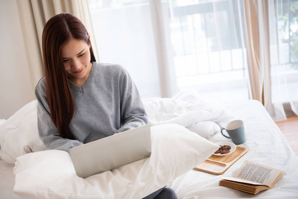 Asian Beautiful woman in grey sweater using laptop while sitting on a bed at home. Relaxing holiday concept. Copy space. Online Shopping. - Foto, afbeelding