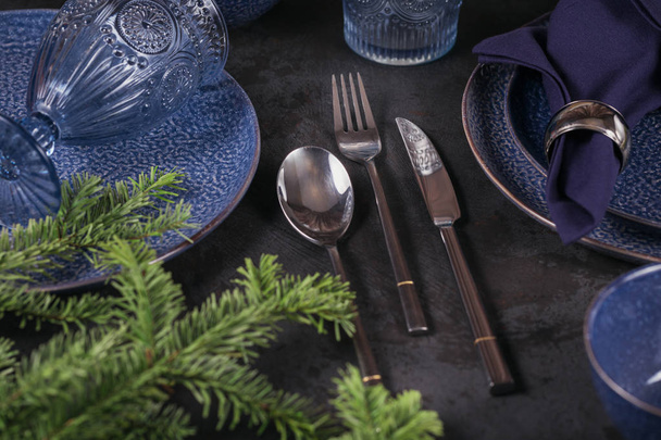Christmas table setting. Dark blue decoration with fir-tree branch. Plates, wine glasses and cutlery with decorative textile on dark background. - Photo, image