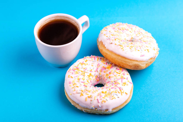 Two delicious vanilla donuts with sprinkle and cup of coffee on bright blue background. Unhealthy, but tasty sweets. Copy space. Horizontal view - Foto, Bild