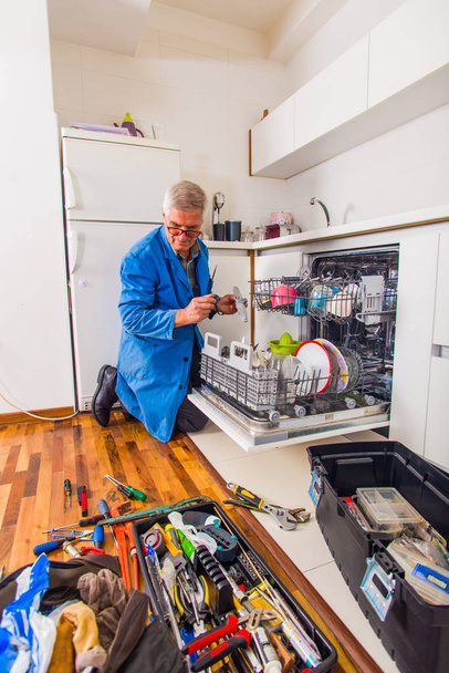 Ώριμος μάστορας ανοίγει το πλυντήριο πιάτων στην κουζίνα με την εργαλειοθήκη κοντά του. - Φωτογραφία, εικόνα
