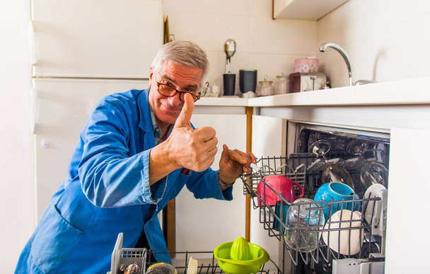 An old age plumber wearing blue uniform and repairing dish washer machine - Foto, afbeelding