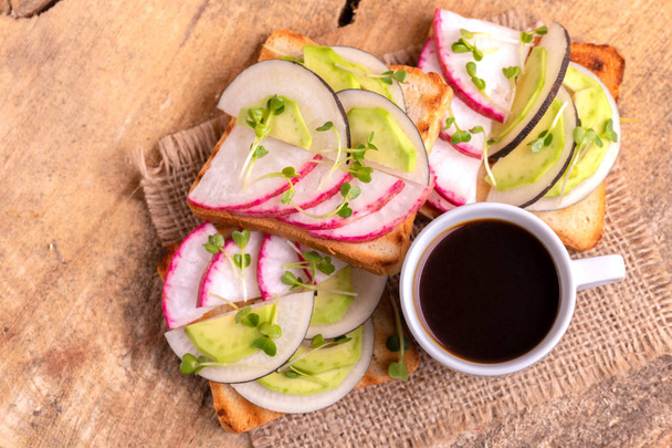 Delicious wheat toasts with radish, avocado and sprouts, cup of strong coffee on burlap napkin. Wooden background. Simple healthy breakfast. Top view - Фото, изображение