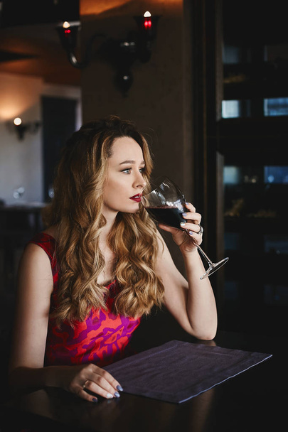 Beautiful and fashionable blonde model girl with sexy red lips in the red-pink dress sits at the table with the glass of red wine in her hand and posing at luxury dark interior - Valokuva, kuva