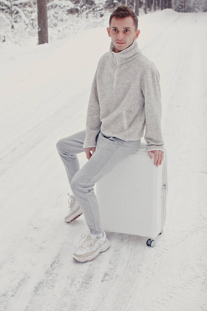 Portrait of young fashionable man in white clothes walking on snow holding suitcase and go pathway pine forest. Winter vacation travel concept. Outdoors in winter. Ski resort, mounting skiing resort - Photo, Image