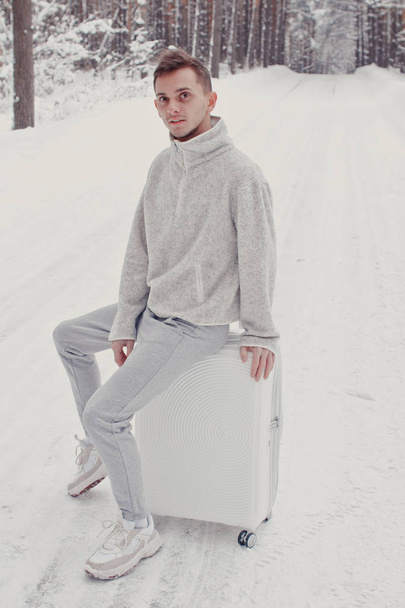 Portrait of young fashionable man in white clothes walking on snow holding suitcase and go pathway pine forest. Winter vacation travel concept. Outdoors in winter. Ski resort, mounting skiing resort - Zdjęcie, obraz