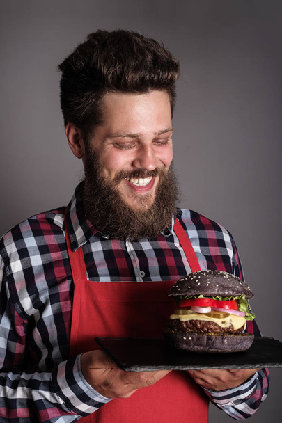 Αρσενικό μάγειρα σε κόκκινη ποδιά δίνοντάς σας φρέσκο αυτο-made μαύρο burger και χαμογελώντας - Φωτογραφία, εικόνα