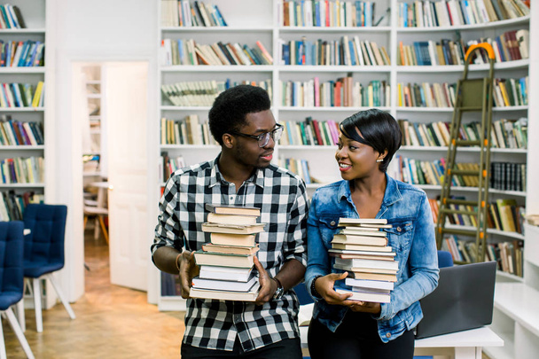 Ευτυχισμένος Αφρικανικός Αμερικανός σπουδαστές αγόρι και κορίτσι σπουδάζει στη βιβλιοθήκη του κρατώντας τα βιβλία και τη διασκέδαση - Φωτογραφία, εικόνα