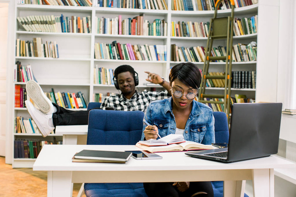 図書館でアフロ ・ アメリカ人学生。女の子は、ラップトップを使用して、音楽に耳を傾け、テーブルの上に彼の足を置く男の背景に本を読んでライブラリ. - 写真・画像