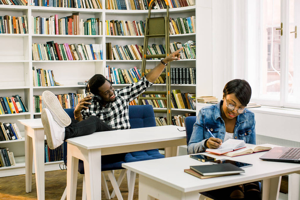 Beautiful African-American students work in the library. The girl smiles and working with the laptop and, reads the books and the boy dances while listening to music. - Foto, Imagem