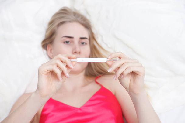 Young woman sitting on a couch, having a fever, holding a thermometer after measuring temperature - Photo, Image