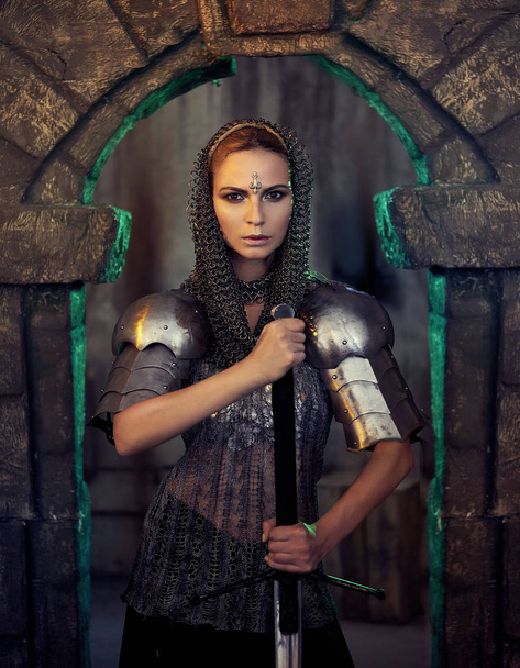 A beautiful warrior girl with a sword and in armor. A model in a chain skirt and a vest in a mysterious plase. Fantasy photo. - Photo, image