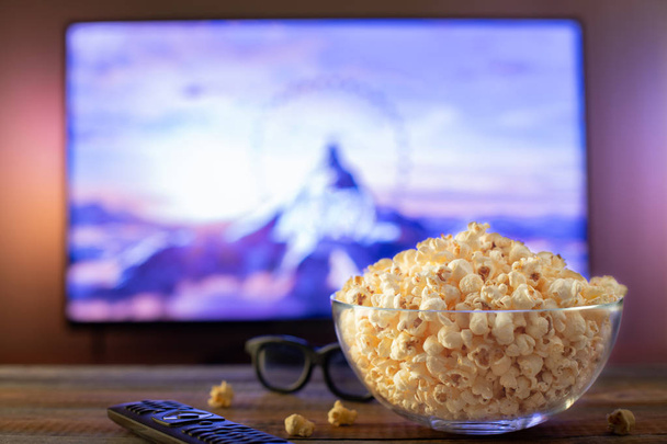 A glass bowl of popcorn, 3d glasses and remote control in the background the TV works. Evening cozy watching a movie or TV series at home - Foto, imagen