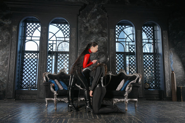 Maîtresse forte maléfique aux cheveux noirs portant une combinaison fétiche en latex et jouant avec son esclave chien en masque de cuir sur fond gothique foncé
 - Photo, image