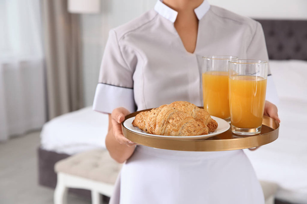 Young chambermaid holding tray with breakfast in bedroom, closeup - Φωτογραφία, εικόνα