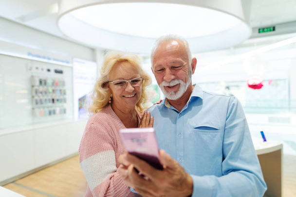 Heureux couple de personnes âgées tester nouveau téléphone intelligent et debout dans le magasin de technologie
. - Photo, image