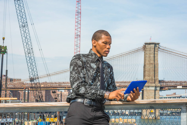 Uomo d'affari afroamericano che viaggia a New York, indossando camicia nera con motivi floreali, cravatta, in piedi al porto, dito che tocca lo schermo, leggendo tablet computer blu. Ponte di Brooklyn sullo sfondo
. - Foto, immagini