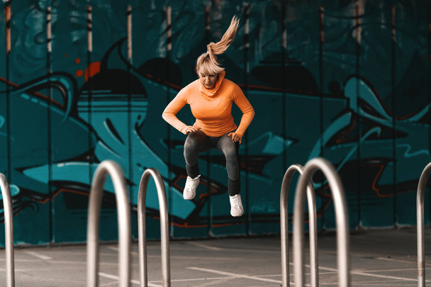 Young beautiful blonde Caucasian woman in sportswear and with ponytail jumping under the bridge. Full length. - Photo, Image