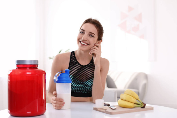 Young woman holding bottle of protein shake at table with ingredients in room - Фото, изображение