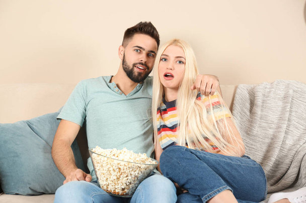 Молодая пара с миской попкорна смотрит телевизор на диване дома
 - Фото, изображение