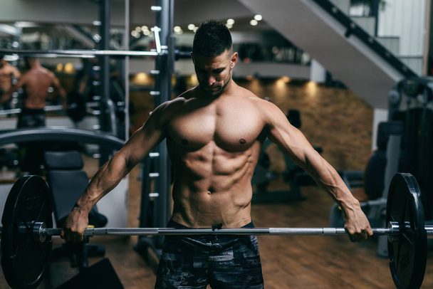 Young strong Caucasian shirtless bodybuilder lifting barbells. Gym interior. - Фото, зображення