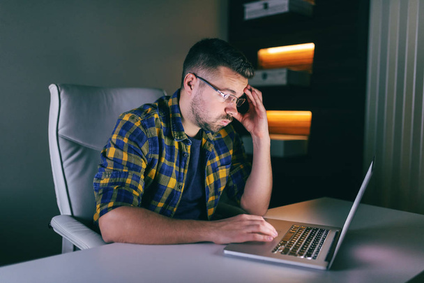 Нещасний бородатий працівник з рукою на голові використовує ноутбук для роботи, сидячи в офісі пізно вночі
. - Фото, зображення