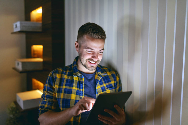 Χαμογελαστός υπάλληλος ντυμένος με καρό πουκάμισο χρησιμοποιώντας tablet κλίνοντας στον τοίχο στο γραφείο του αργά τη νύχτα. - Φωτογραφία, εικόνα