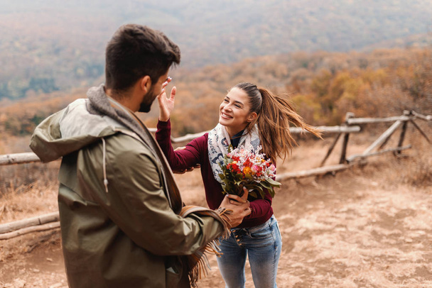 Mixed race man giving bouquet to his beautiful girlfriend while standing on the glade at autumn time. In background forest. - Photo, Image