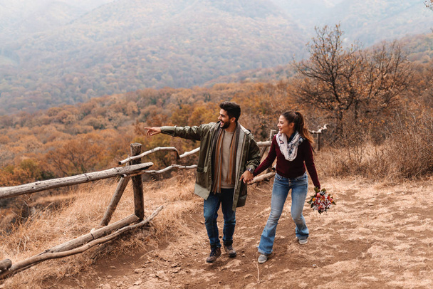 Couple in love walking in the nature at autumn and holding hands. Man pointing at view while woman holding bouquet in hand. In background forest and hills. - Foto, afbeelding