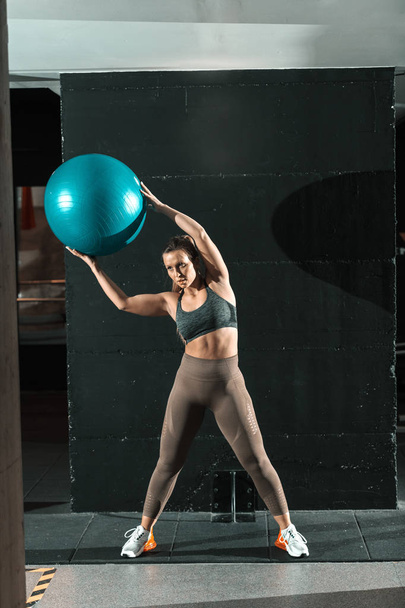 Σπορ εστιασμένη σπορ καυκάσιος γυναίκα ντυμένη σε αθλητικά είδη και με αλογοουρά άρση μπάλα pilates ενώ στέκεται στο γυμναστήριο. - Φωτογραφία, εικόνα