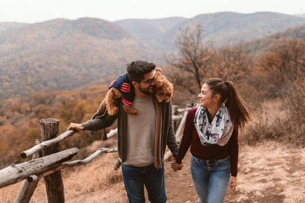 Happy couple in love holding hands and taking a walk in the nature at autumn. Man having apricot poodle on his shoulders. - Photo, Image