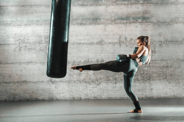 Kaukaski kobieta w sportowej i rękawice bokserskie, kopiąc torba na siłowni. Pełnej długości. Ściany w tle. - Zdjęcie, obraz