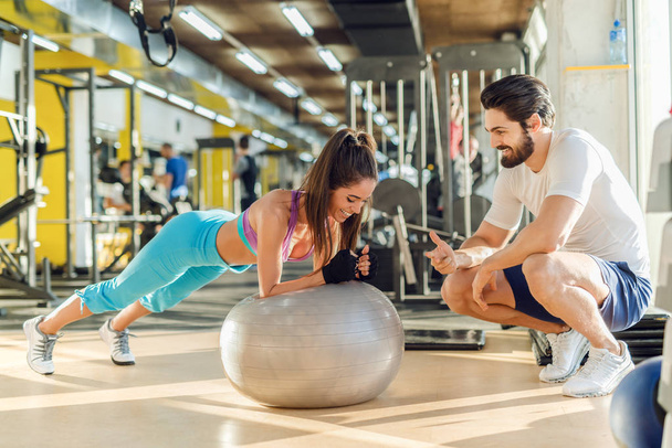Sporty smiling woman doing planks on pilates ball while her personal trainer crouching next to her and cheering for her. - Photo, Image
