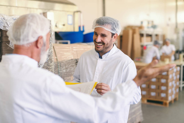 Два кавказских рабочих в защитном костюме разговаривают и улыбаются стоя на пищевой фабрике
. - Фото, изображение