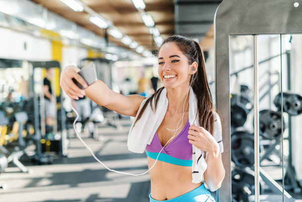 Femme caucasienne avec serviette autour du cou et écouteurs dans les oreilles souriant et prenant selfie tout en se tenant dans une salle de gym
. - Photo, image