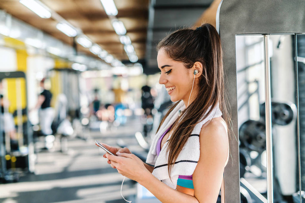 Κοντινό πλάνο της χαμογελώντας καυκάσιος γυναίκα επιλογή μουσικής στο smart phone ενώ στέκεται σε ένα γυμναστήριο. Πετσέτα γύρω από το λαιμό. - Φωτογραφία, εικόνα