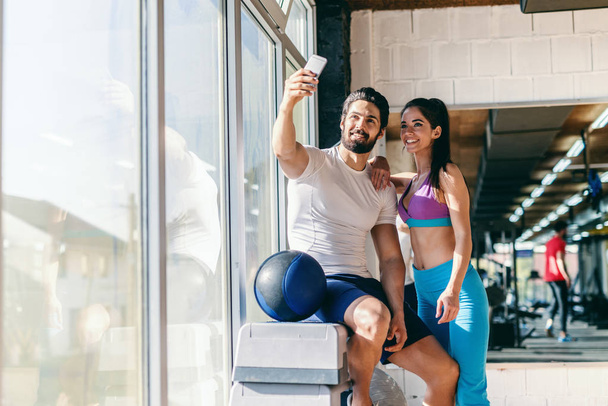 Linda pareja deportiva tomando selfie con teléfono inteligente en el gimnasio
. - Foto, Imagen
