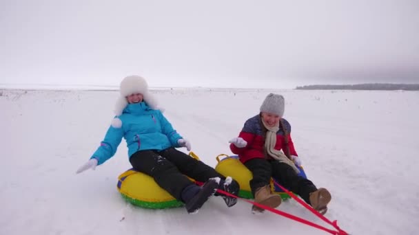 Zabawny matek i ich córka są jazda snowy płycie wzdłuż drogi mroźną zimę, śmiejąc się i grając śnieżkami. Życie rodzinne jako sposób życia. Dziewczyny śmieją się, grać śnieżkami i jeździć na sankach - Materiał filmowy, wideo