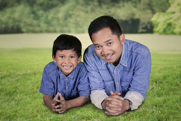 Close up of young man smiling at the camera while lying with his son on the green grass. Shot in the park - Foto, afbeelding