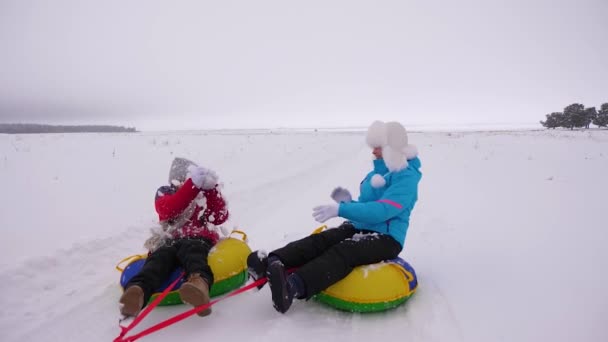 Funny mothers and their daughter are riding snowy plate along snowy winter road, laughing and playing snowballs. Family life as way of life. Girls laugh, play snowballs and ride on sled - Footage, Video