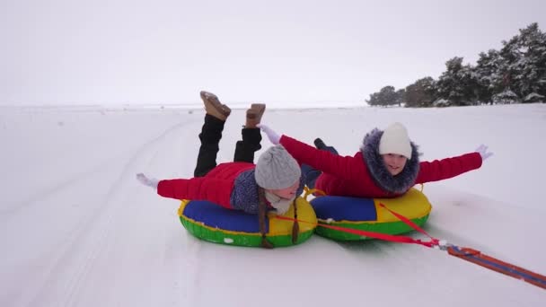 children in red jackets ride through snow in winter on an inflatable snow pipe and play super heroes. happy girls relaxing in the winter park for christmas holidays. Slow motion - Footage, Video