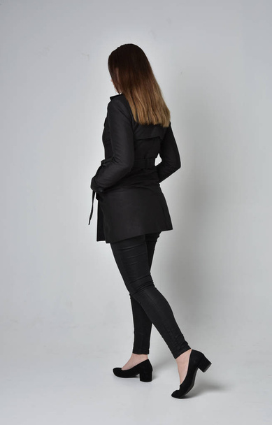 full length portrait of a brunette girl wearing long black coat, standing pose with back to the camera on grey studio background. - Fotó, kép