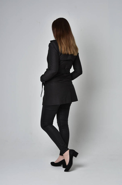 full length portrait of a brunette girl wearing long black coat, standing pose with back to the camera on grey studio background. - Fotografie, Obrázek