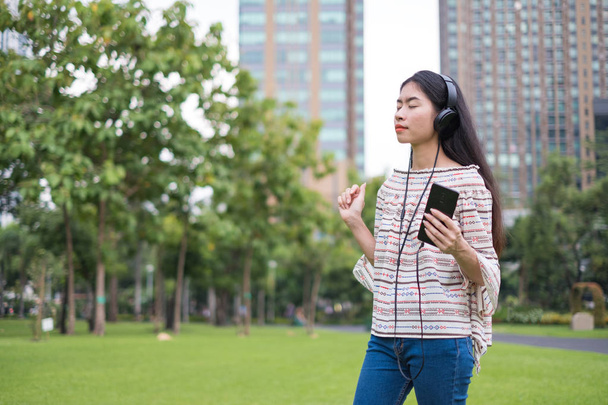 Asian young girl stands listening to music from a smartphone using headphones. At public park the city center happy and relax On his vacation. she is 25-30 years old. - Valokuva, kuva