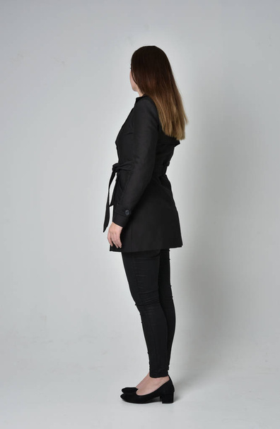 Pełna długość Portret brunetka Girl ubrany długi czarny płaszcz, stojący poza z powrotem do aparatu na studio szary tło. - Zdjęcie, obraz