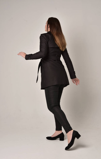full length portrait of a brunette girl wearing long black coat, standing pose with back to the camera on grey studio background. - Foto, immagini