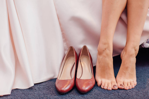 Γυμνά πόδια του μια νύφη που καθόταν δίπλα τα παπούτσια της και το φόρεμα κατά τις προετοιμασίες του γάμου - Φωτογραφία, εικόνα