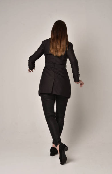 Pełna długość Portret brunetka Girl ubrany długi czarny płaszcz, stojący poza z powrotem do aparatu na studio szary tło. - Zdjęcie, obraz